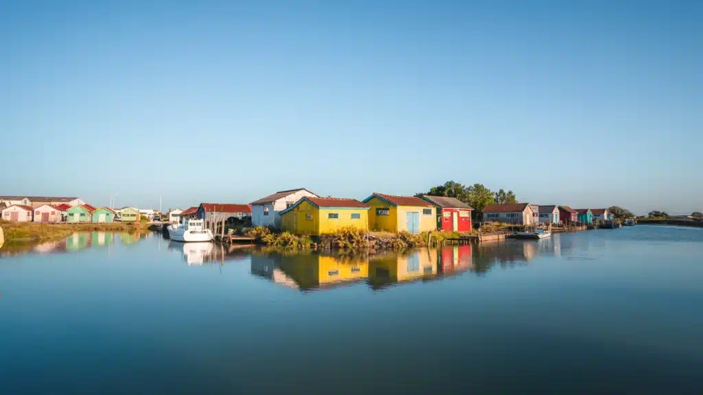 Rangée de petites maisons colorées au bord d’un canal calme, reflétées dans l’eau sous un ciel bleu.