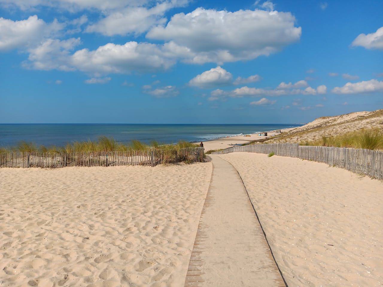 Chemin en bois sur une plage de sable bordée de dunes, clôtures en roseaux, mer bleue et ciel nuageux.
